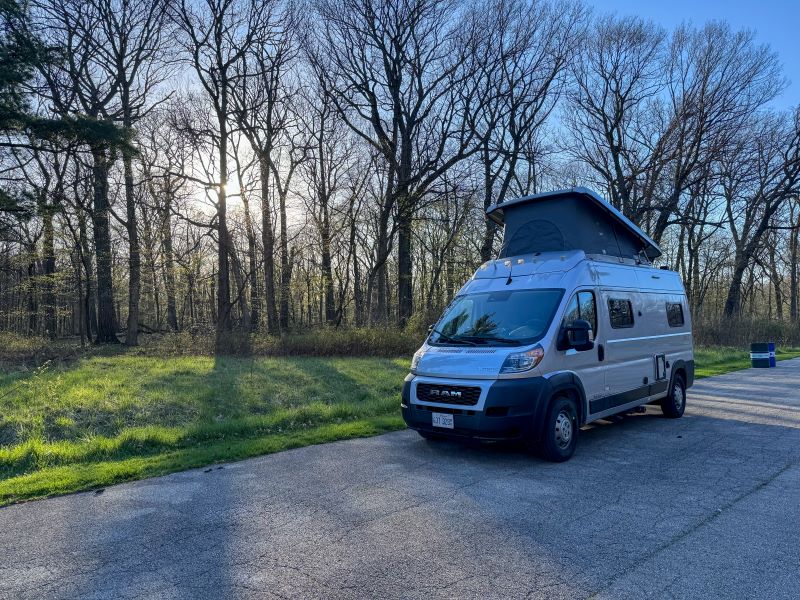 Light gray camper van with pop-top parked on asphalt next to a sunny forest. Perfect for an rv rental trip.