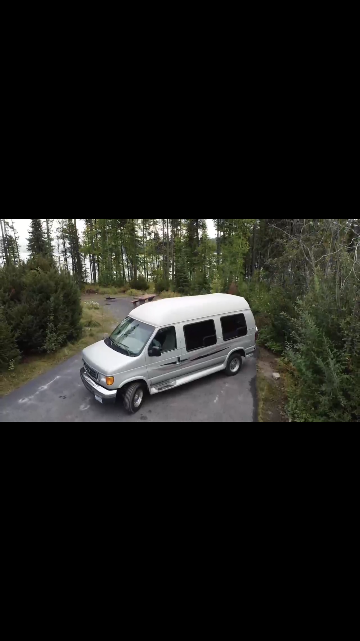An RV rental silver van awaits adventure at a tranquil forest campsite with a lake in the background.