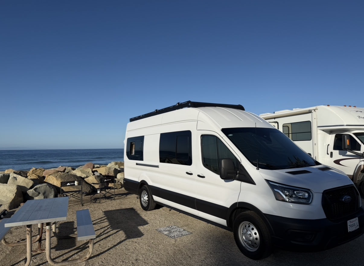 White Ford Transit rv rental camper van parked oceanfront. Sunny day, beach setup with picnic table and chairs.