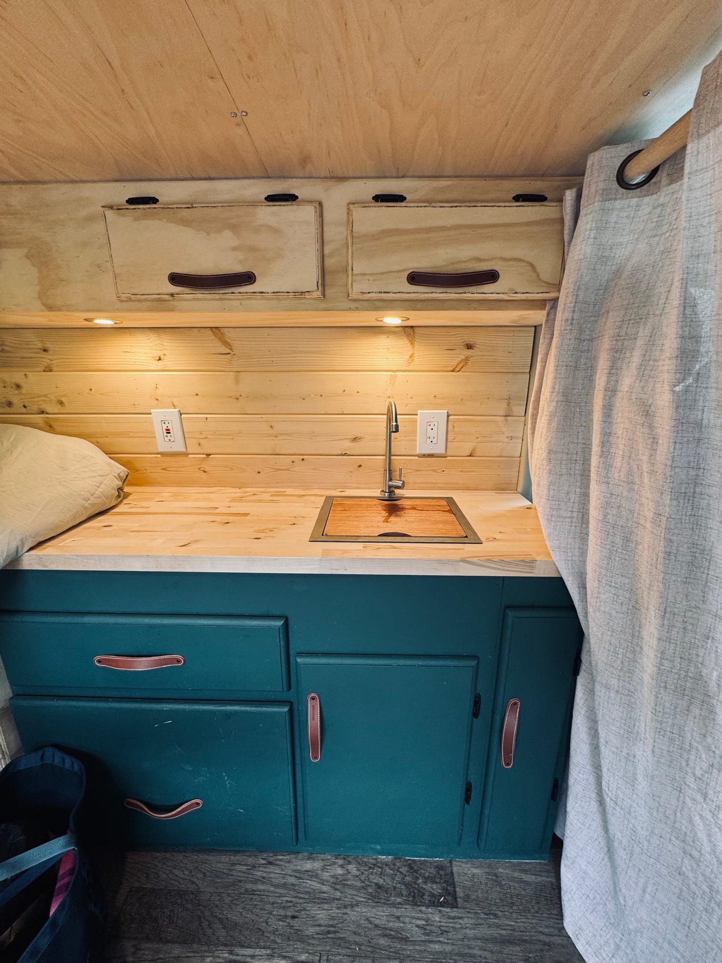 Inside a modern RV rental: a cozy kitchenette with wood counters, dark green cabinets, sink, and overhead storage.