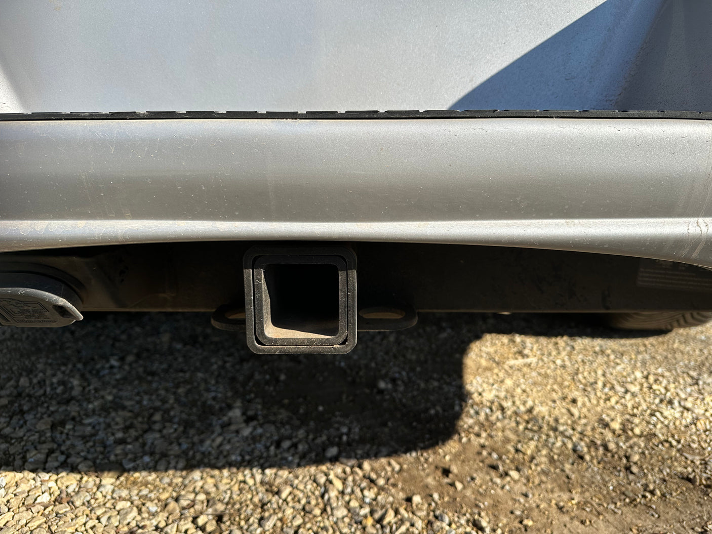 Silver RV rental's rear with trailer hitch receiver and electrical connection for towing, on gravel.