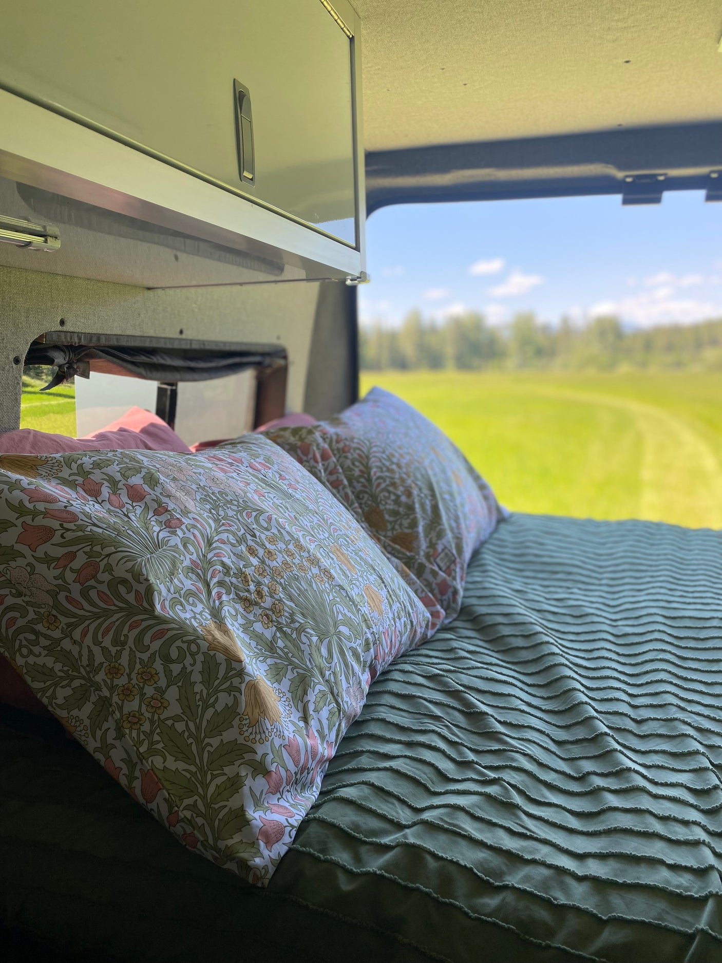 Cozy RV rental bed with patterned pillows, a textured green comforter, and a stunning view of a bright green field.