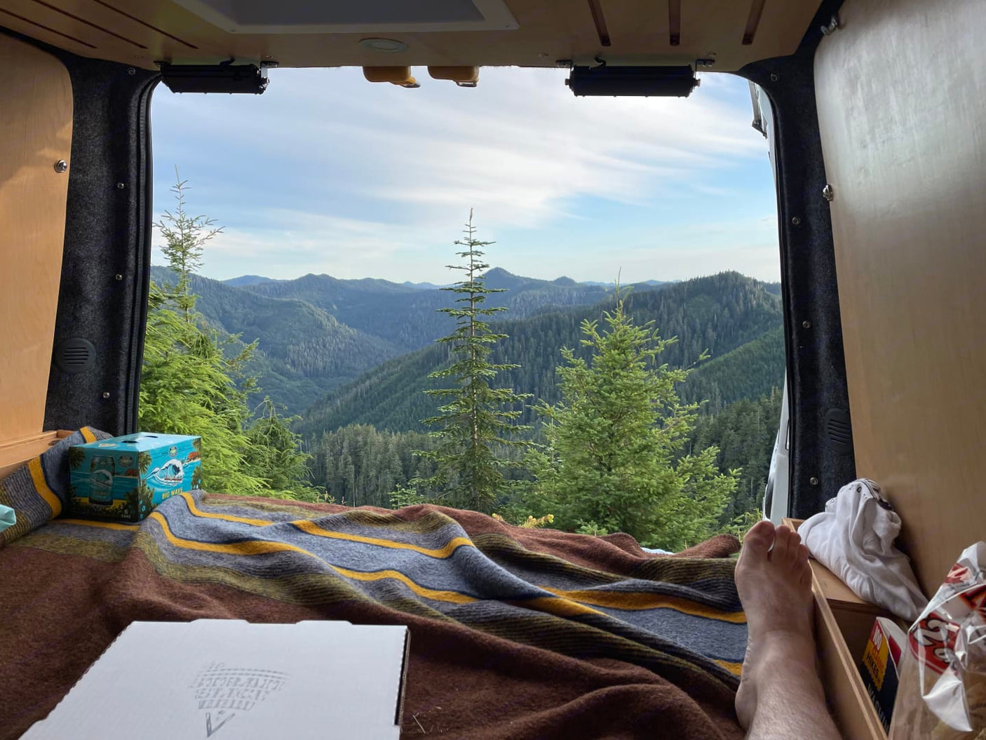 Legs propped, enjoying a breathtaking forest mountain view from the open rear of an rv rental van. Pizza & drinks present.