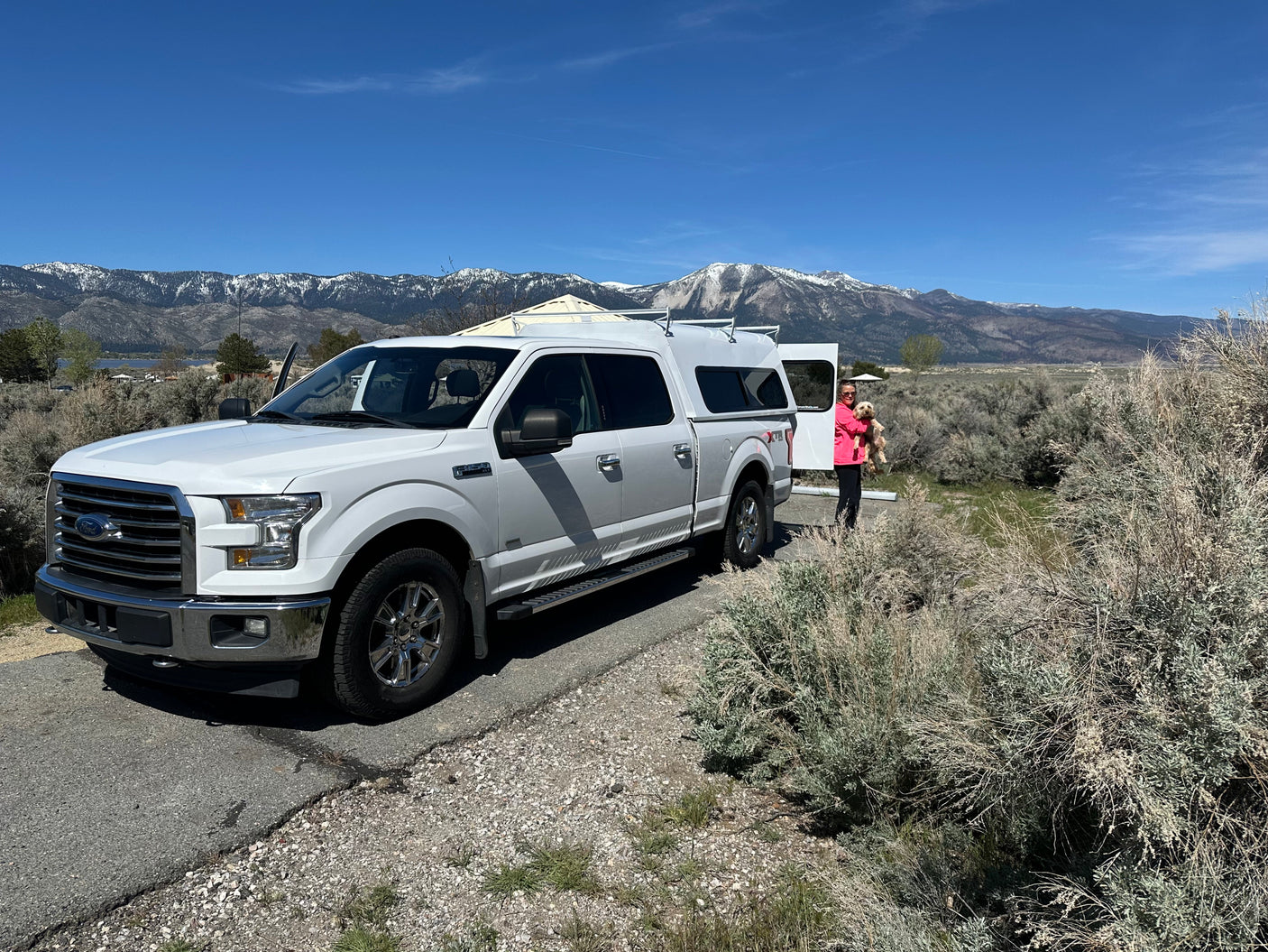 An RV rental journey: White truck with camper, a person holding a dog, scenic snowy mountains, and a lake.