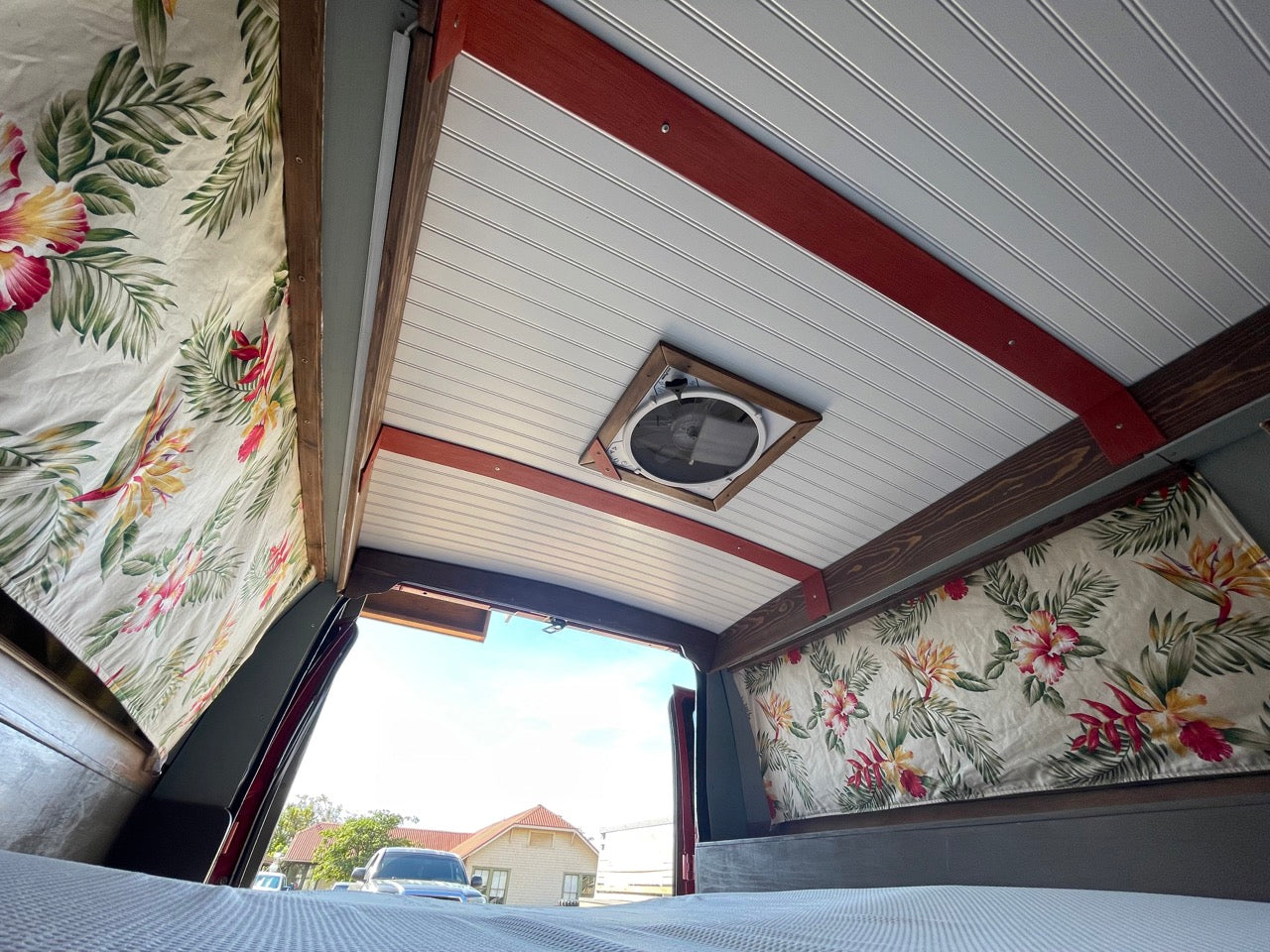 Cozy RV rental interior: tropical curtains, wood ceiling, bed, and fan visible in this custom van conversion.