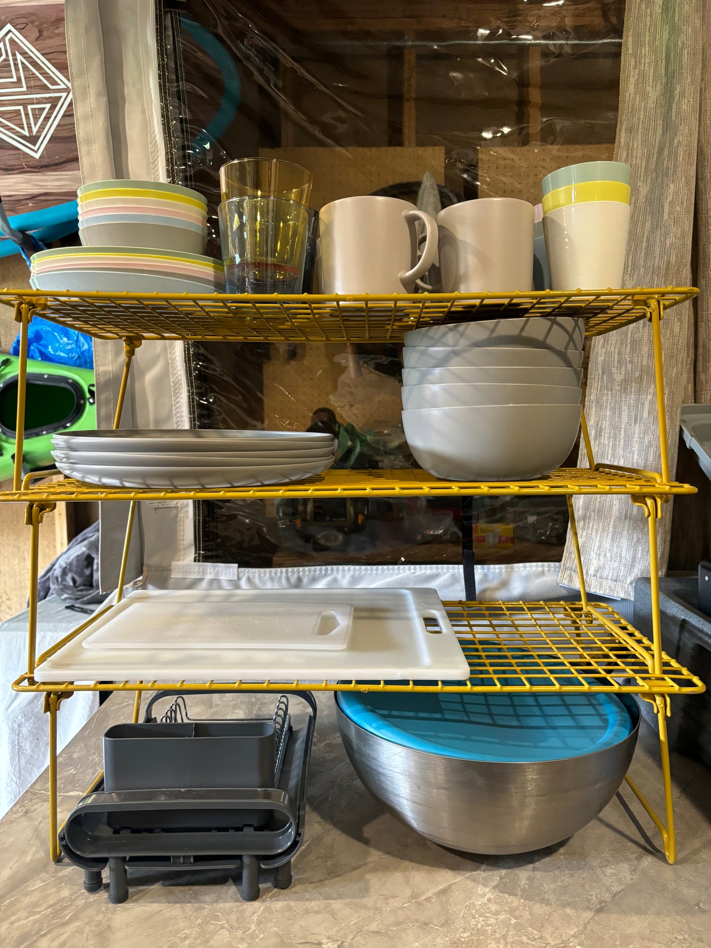 Efficient RV rental kitchen: yellow shelves hold stacked colorful dishes, mugs, and a drying rack with cutting boards.
