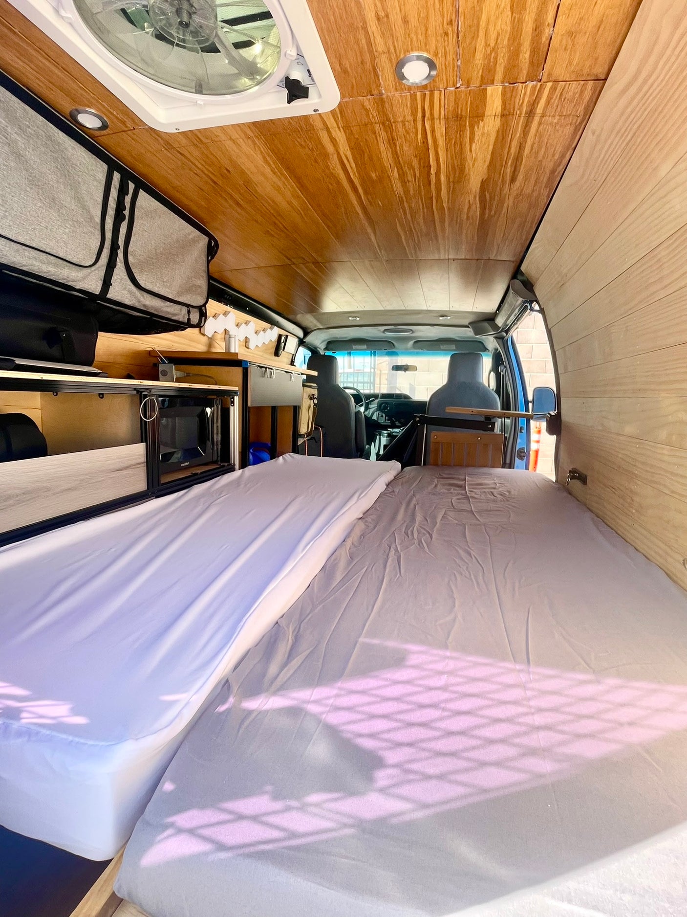 Inside an RV rental van: Twin beds, wood paneling, overhead storage, and a small kitchen unit with a microwave.