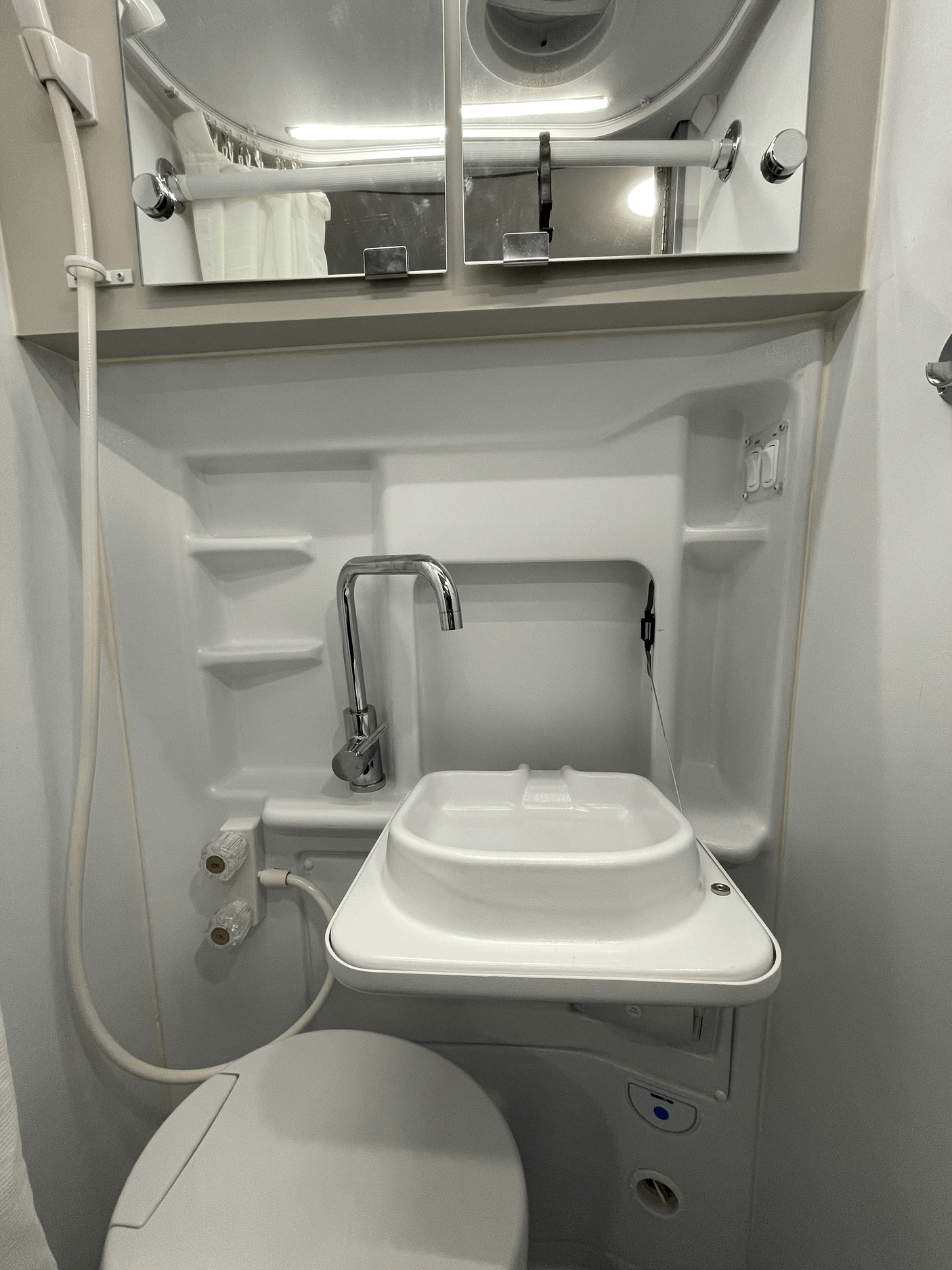Inside a compact RV rental bathroom: features a toilet, fold-down sink, and shower hose with mirrored cabinet.