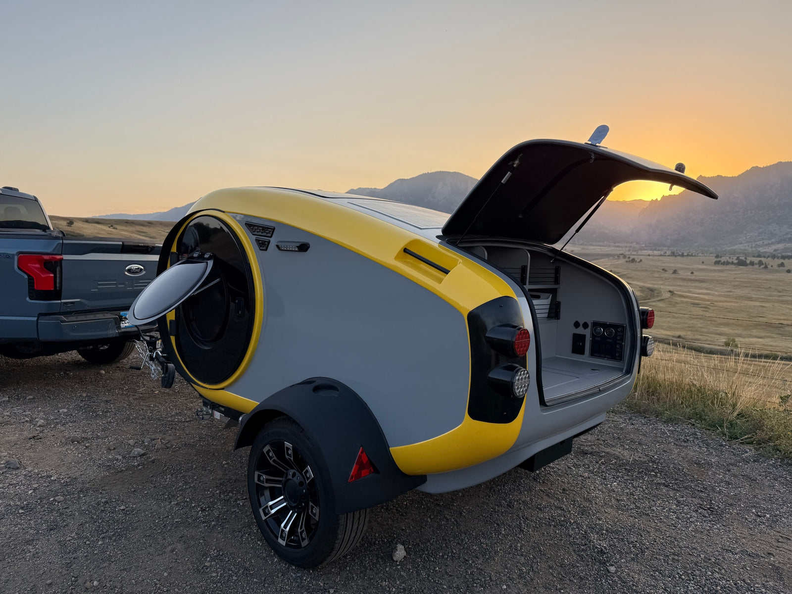 Modern RV rental teardrop camper with open rear, hitched to an electric truck. Scenic sunset over mountains for outdoor travel.