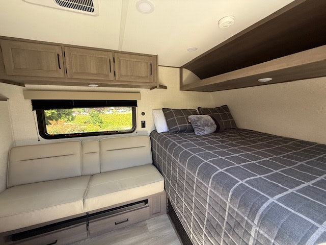 Bright RV rental interior featuring a cozy bedroom area with plaid bedding, comfortable seating, window views, & ample storage.