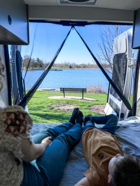 Experience RV rental comfort: Two people relax on a bed inside a van, gazing at a serene lake through the mesh screen.