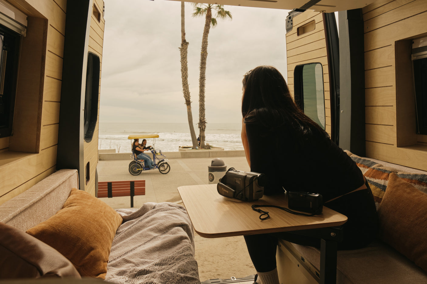Serene beach view from an RV rental's open door. A woman watches a pedal bike go by, cameras ready for coastal memories.