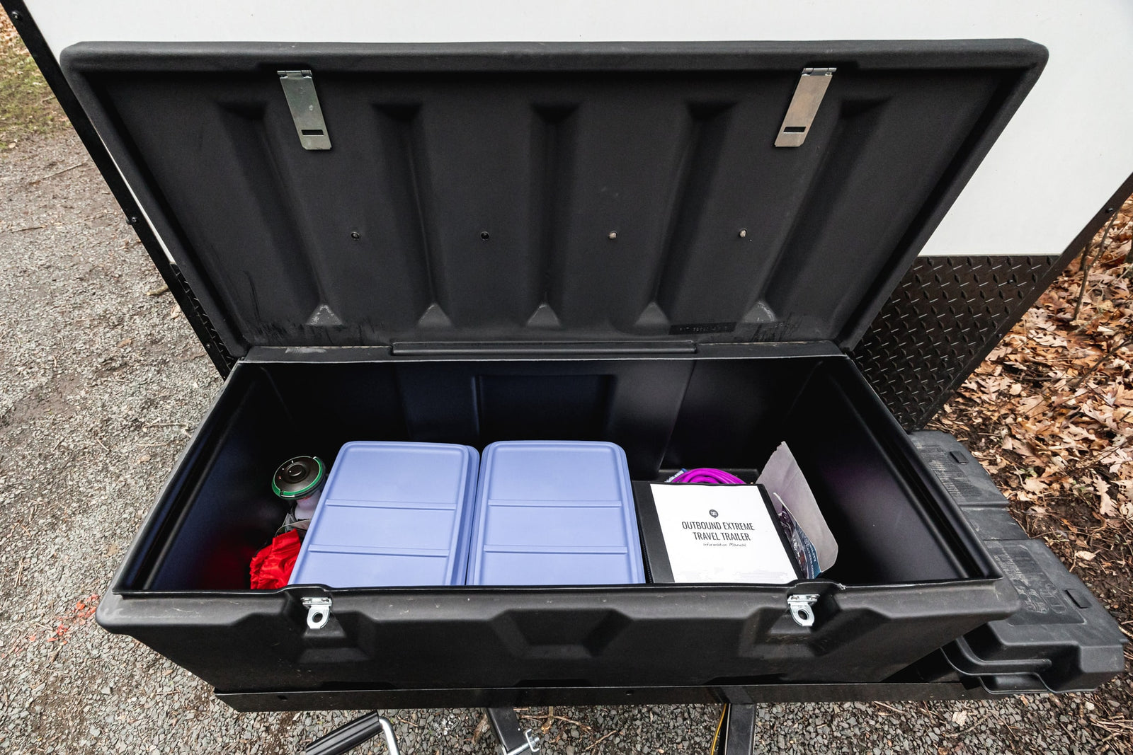 Opened front storage box on an RV rental travel trailer, neatly organized with blue containers, a red bag, and manual.