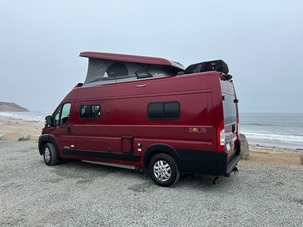 Red Solis rv rental camper van with pop-top roof, parked on gravel by the ocean under an overcast sky.
