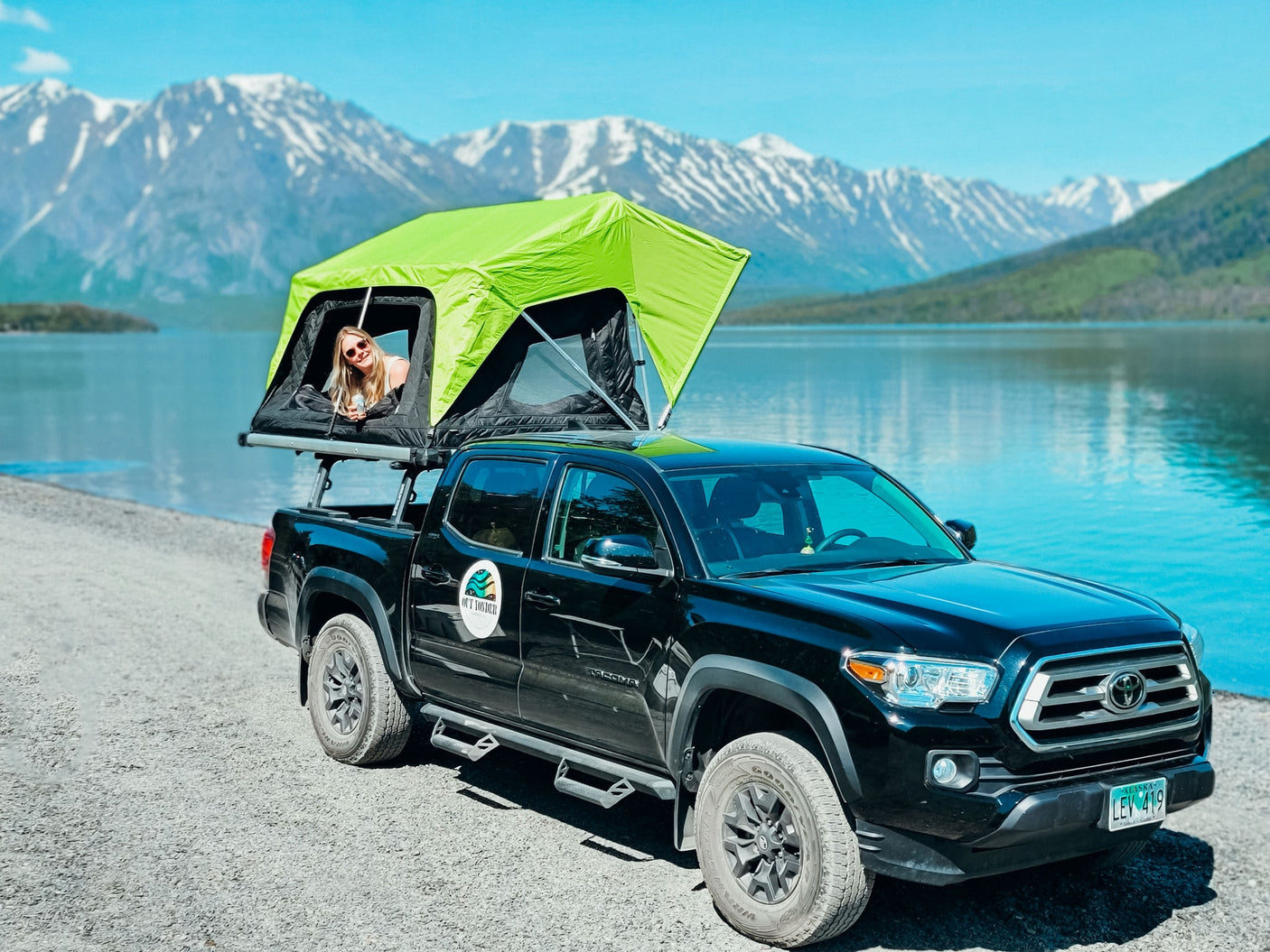 Experience adventure with our RV rental! A woman relaxes in a bright green rooftop tent on a black Toyota Tacoma by a scenic lake with mountains.
