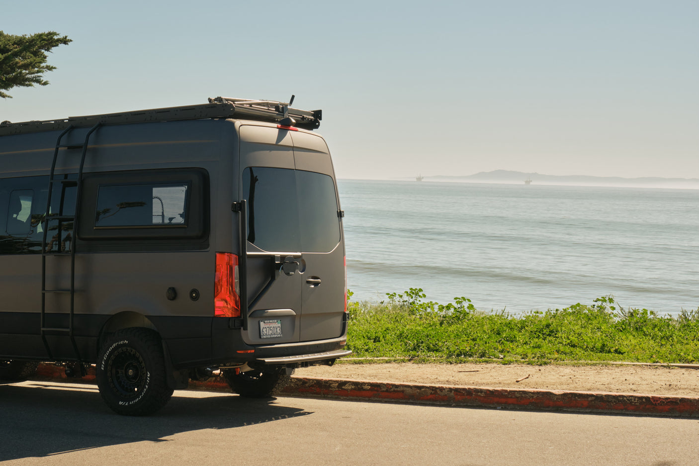 RV rental ready: A grey camper van with all-terrain tires parked on a sunny coastal road, ocean view & distant rigs.