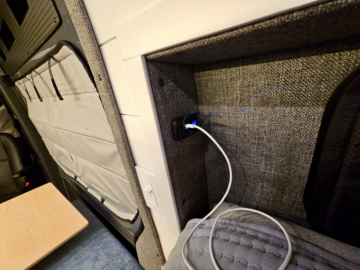 RV rental charging nook: Fabric-lined cubby with illuminated USB port & white cable. Bed, table & pleated window shade for comfort.