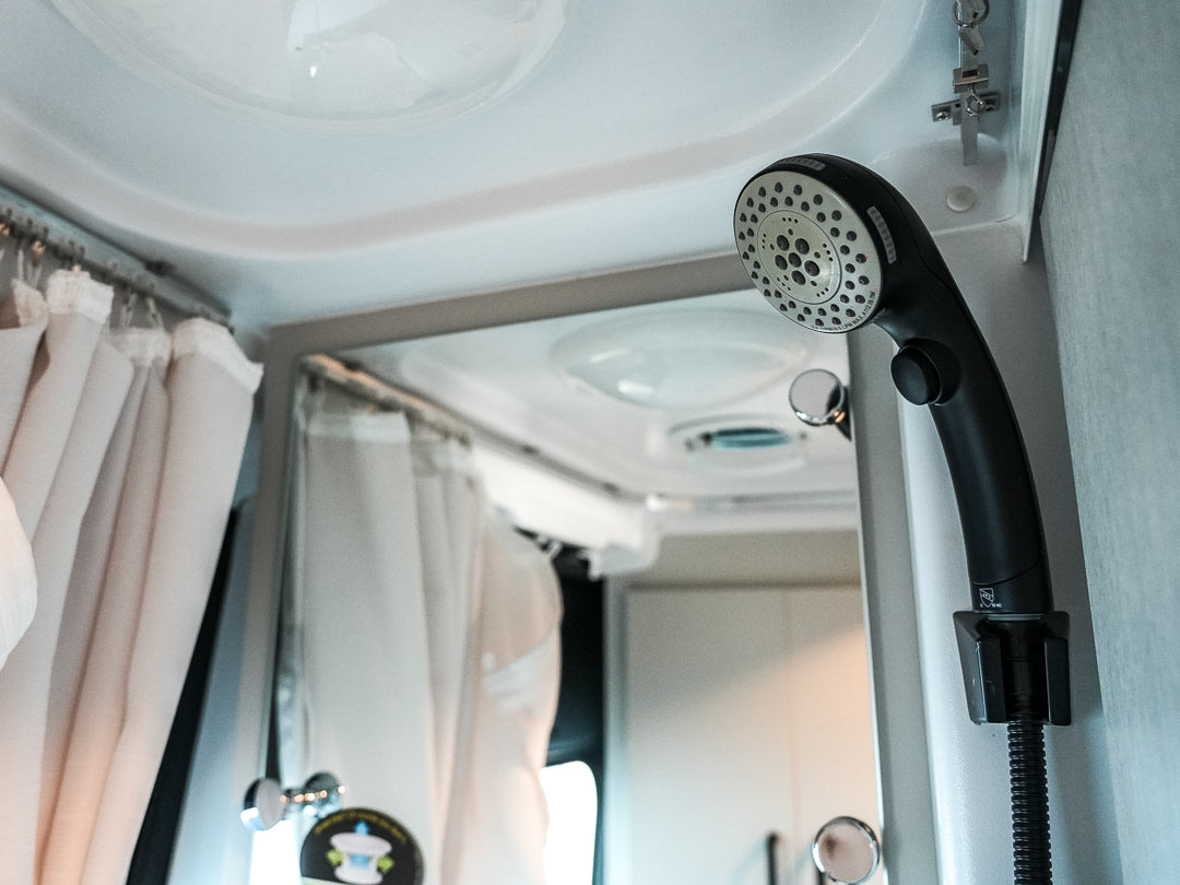 Adjustable black showerhead in a compact RV rental bathroom, with light privacy curtains and reflective surfaces.