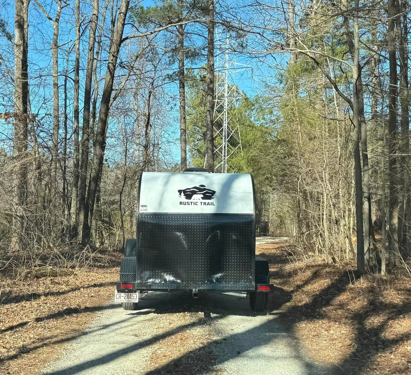 Rear view of a Rustic Trail camping trailer on a wooded path, perfect for an RV rental outdoor adventure.
