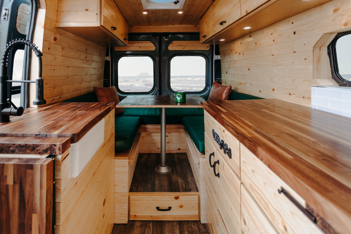 Cozy rv rental camper van interior. Features include wood paneling, a kitchenette, dinette with green cushions, and ocean views.