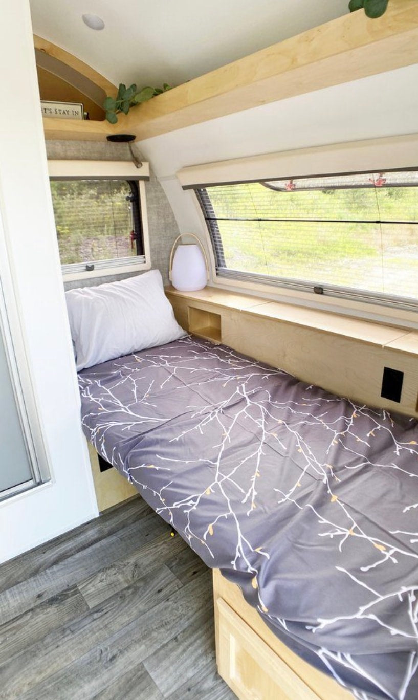 Inside a compact RV rental: a comfortable single bed with branch-patterned sheets, window views, and light wood interior.