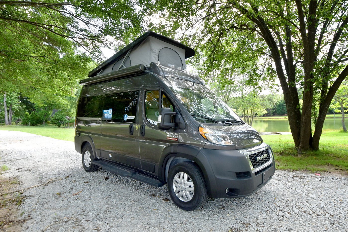 Modern dark grey RV rental camper van with pop-top roof on a gravel path, amidst green trees and a serene lake. — photo 1