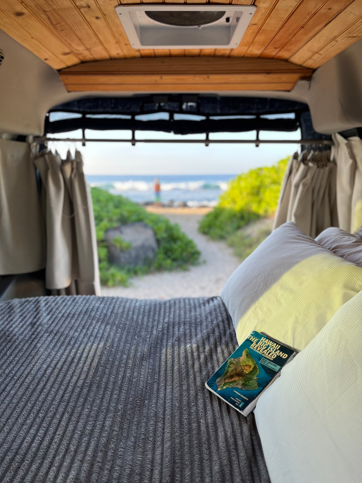 Relaxing inside an RV rental. A Hawaii guidebook rests on the bed, with a serene beach view beyond.