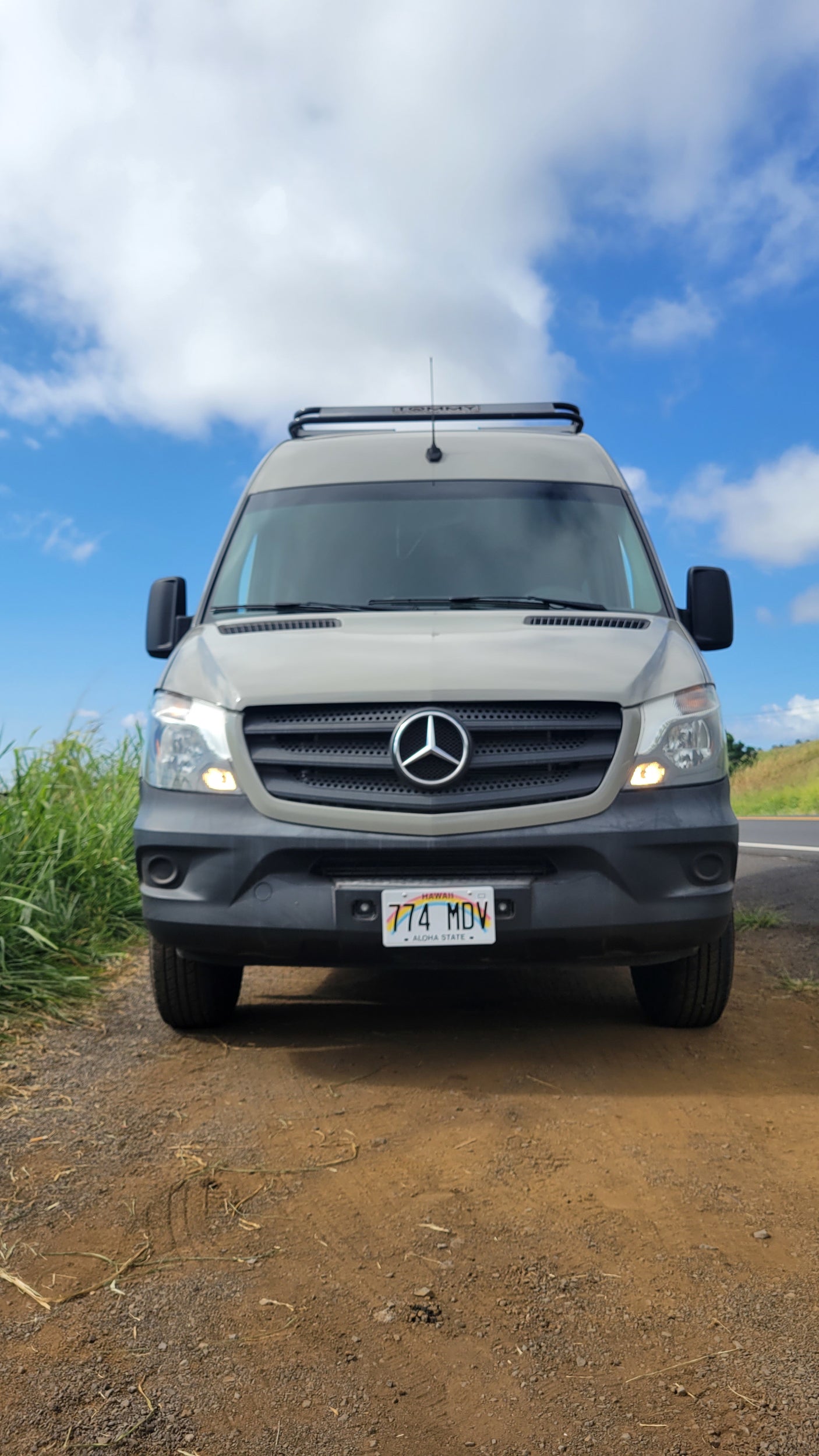 RV rental: Front view of a gray Mercedes-Benz Sprinter camper van parked on a dirt shoulder in Hawaii.