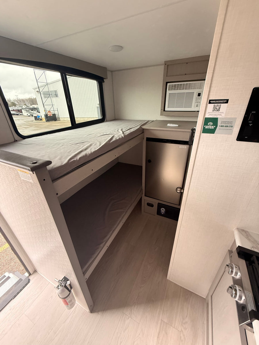 Interior of an RV rental features stacked bunk beds, mini-fridge, AC unit, and a glimpse of a stove. Light-toned, functional space.