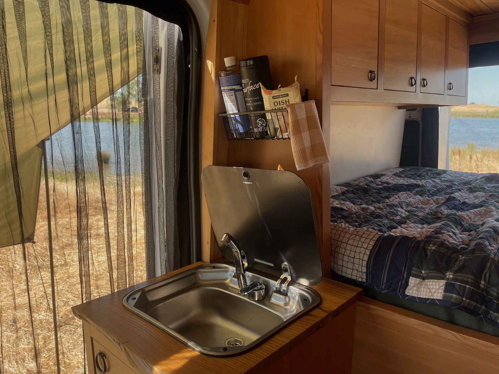 RV rental interior: compact kitchen with sink & supplies, cozy bed. A mesh window reveals a serene lake and wild landscape.