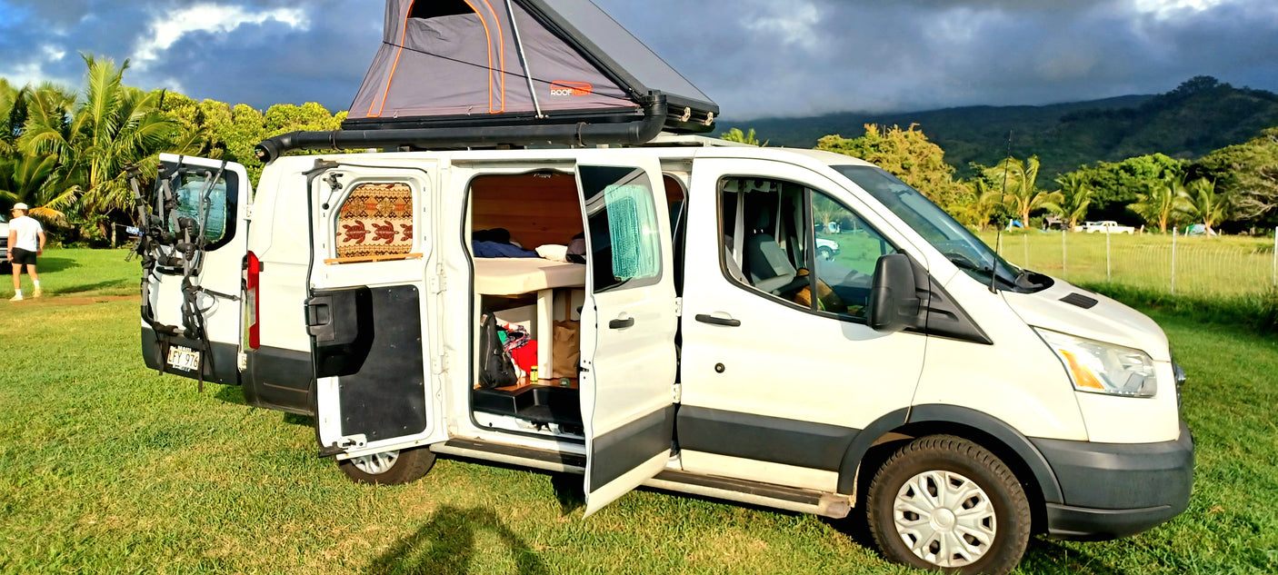 An RV rental Ford Transit camper van with open doors, showing its cozy bed & rooftop tent, parked on a grassy field in Maui.