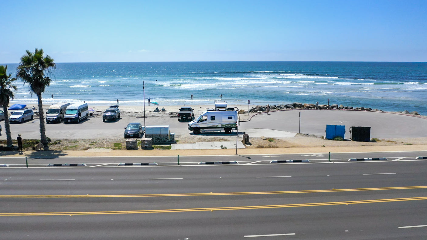 RV rentals parked at a sunny beachside lot. Waves crash as people enjoy the ocean view and sand.