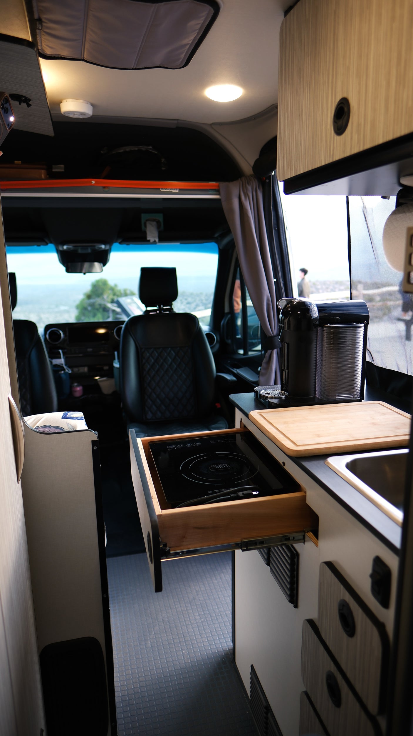 Inside a modern RV rental: kitchenette with induction cooktop, sink, Nespresso, and front cabin view of a scenic landscape.
