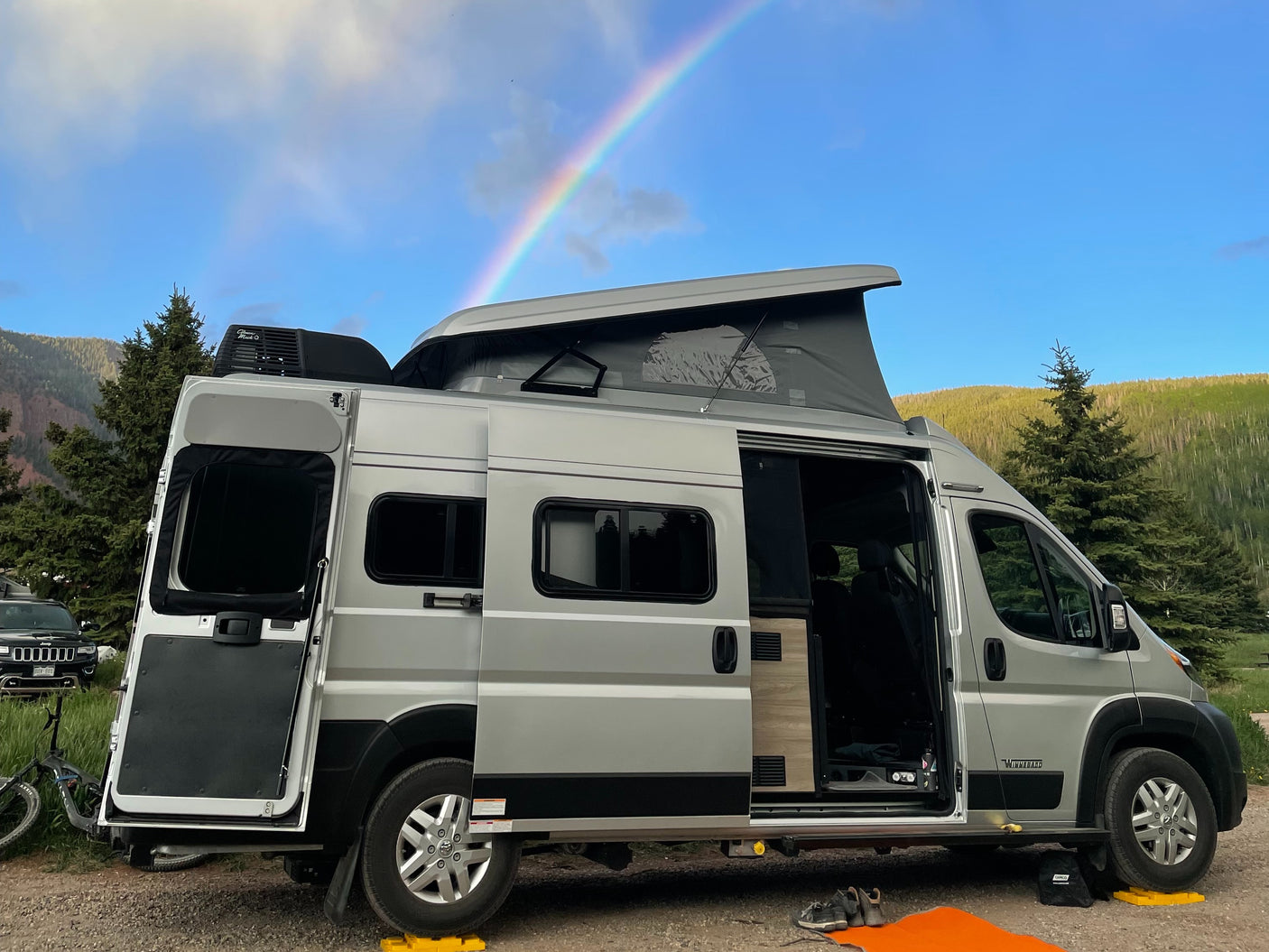 Enjoying nature in an RV rental! Camper van with pop-top under a rainbow, parked among trees and mountains.