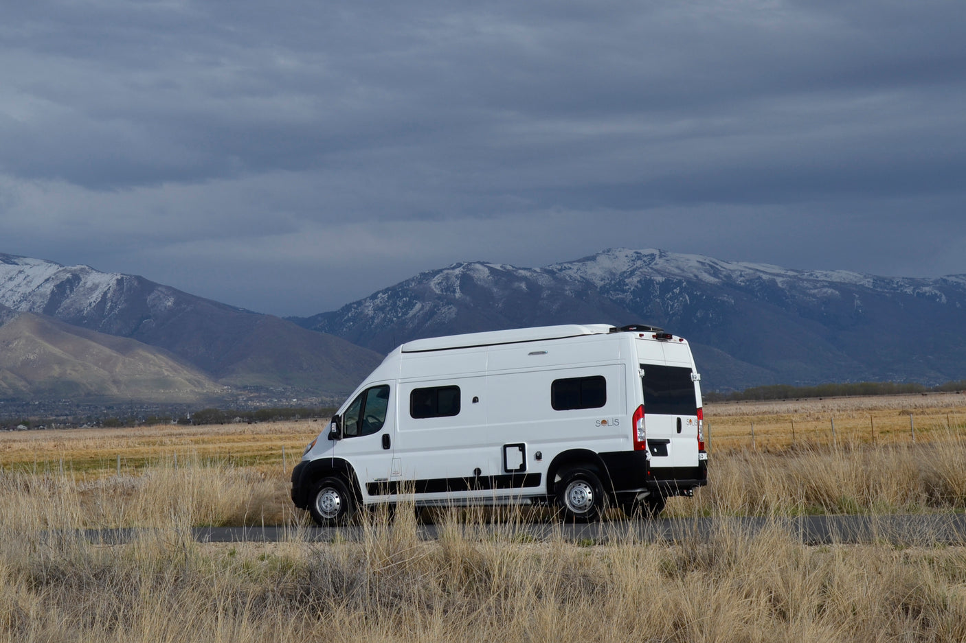 A white RV rental camper van is parked on a road in a dry field, with majestic snow-capped mountains under cloudy skies. Ideal for travel.