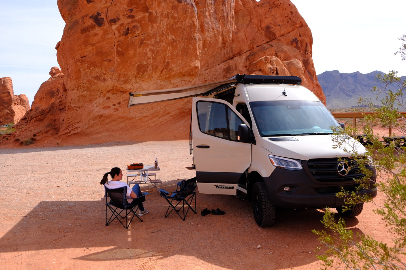 Desert adventure with an rv rental. Campervan parked by red rocks, woman relaxing under awning, enjoying the view.