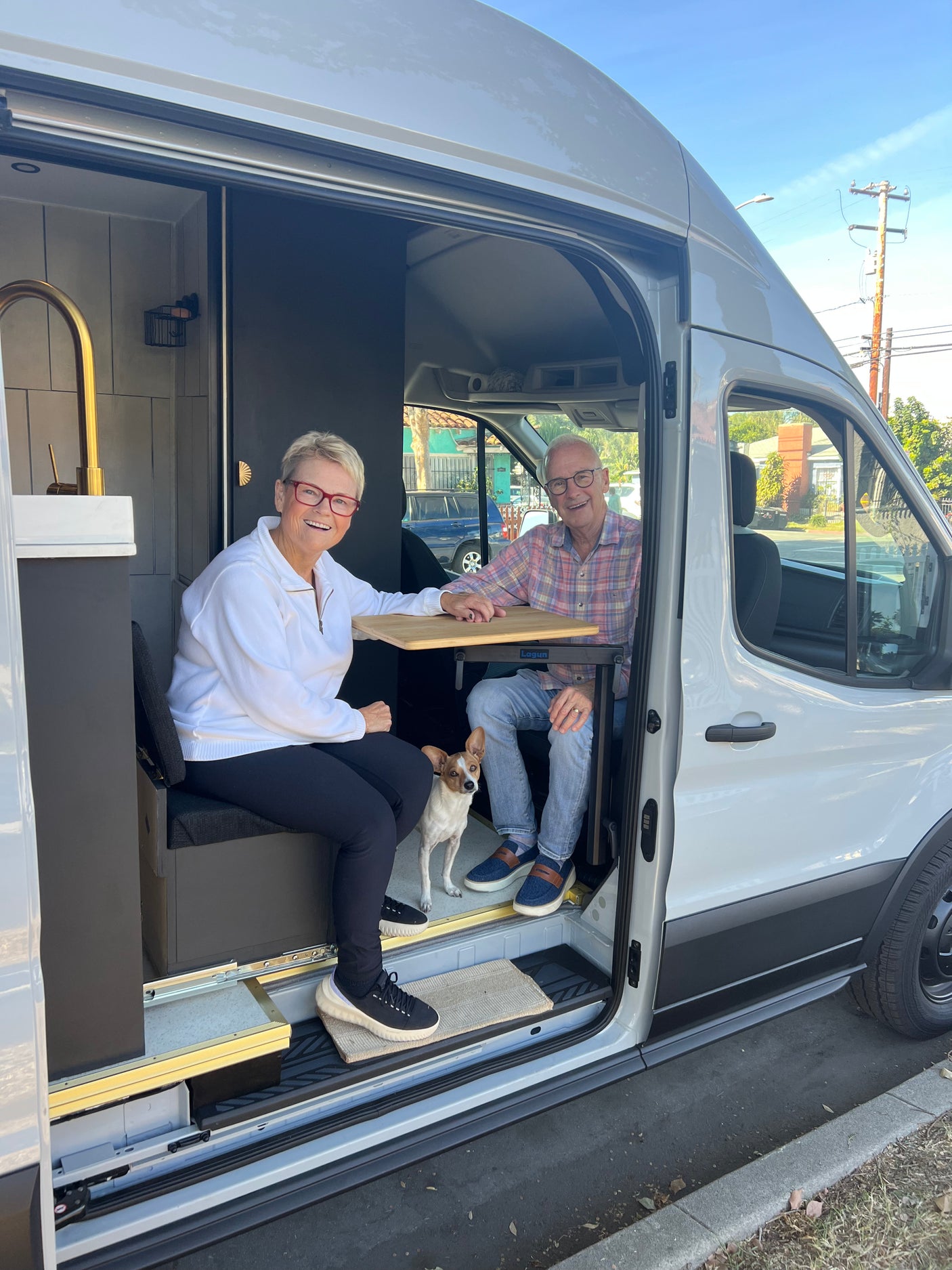 Inside a bright RV rental van, a smiling couple and their dog relax at a table, ready for travel.