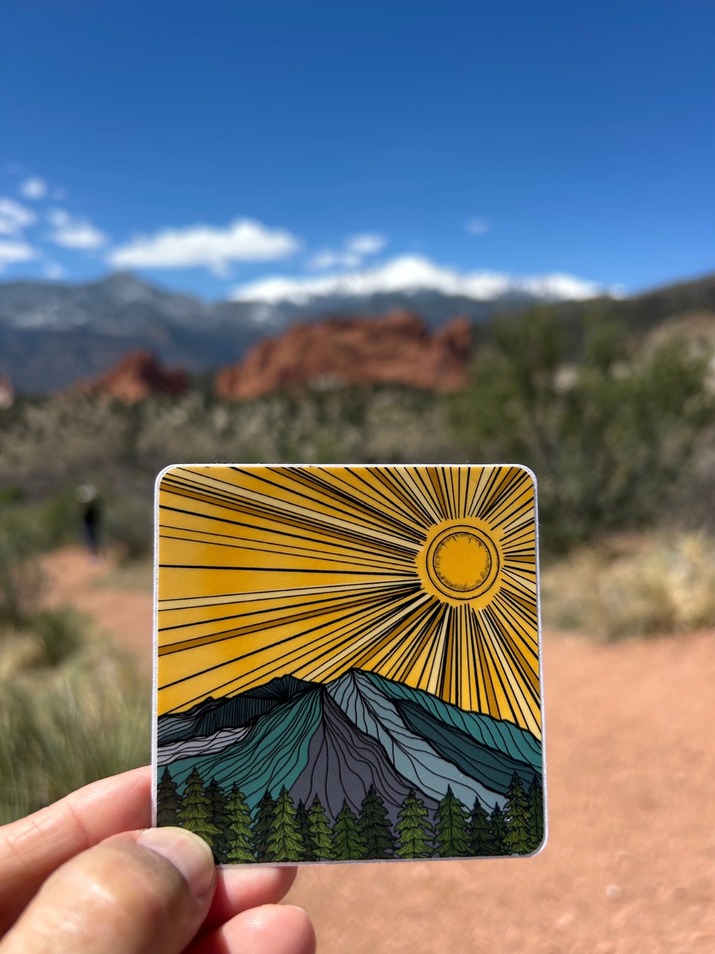 Hand holds a mountain & sun sticker, mirroring a blurred scenic mountain backdrop. Great for your next rv rental adventure!