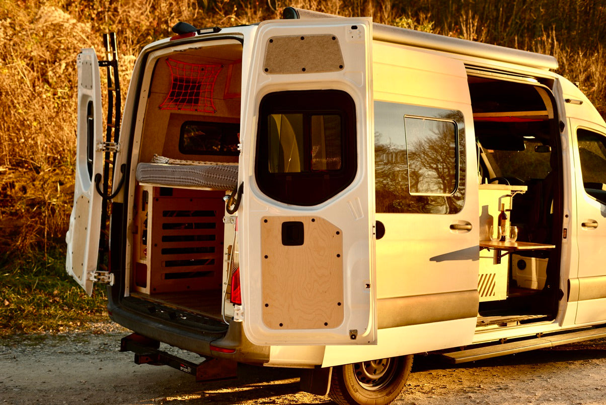Spacious white camper van RV rental, rear and side doors open. Inside: bed, storage, table with wine. Ready for outdoor adventure.