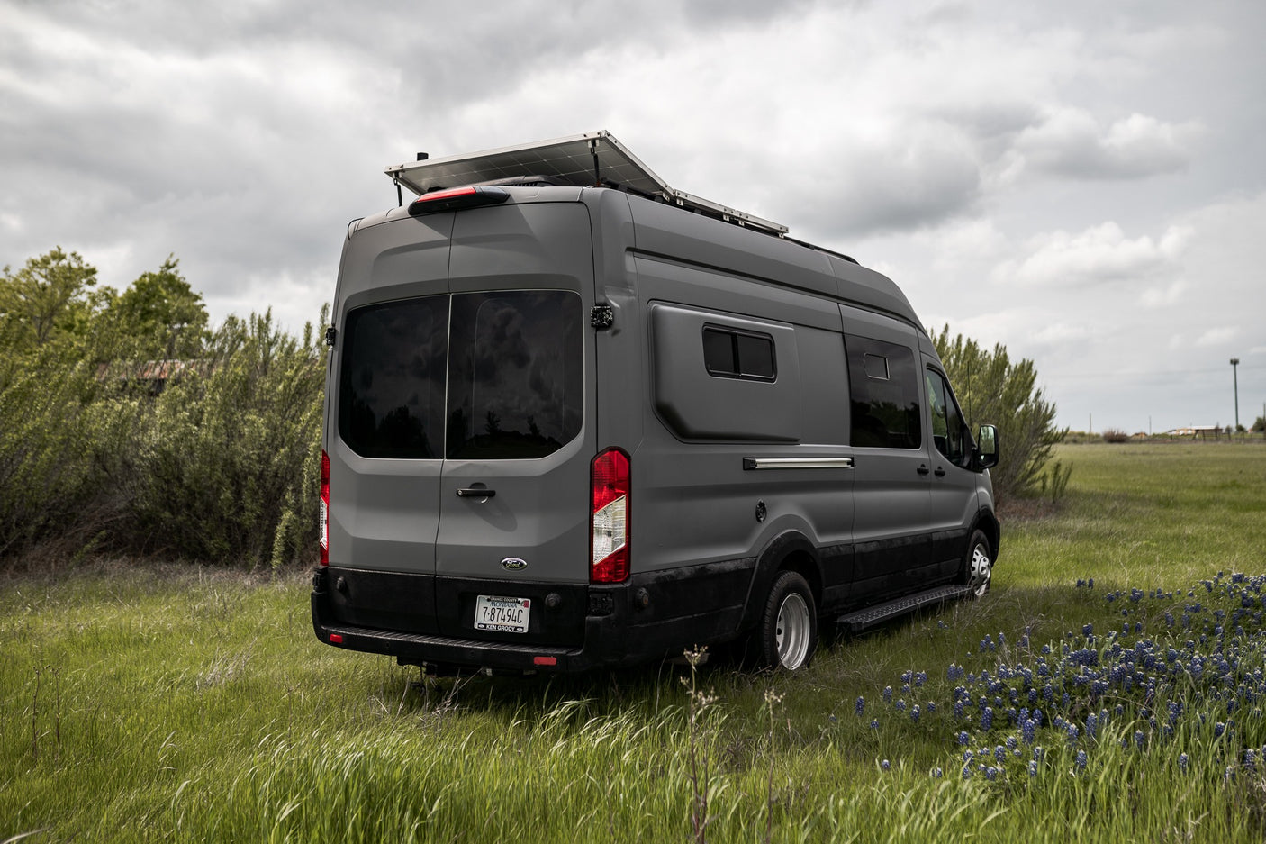 Gray Ford Transit camper van (RV rental) with solar panels parked in a grassy field with bluebonnets under a cloudy sky.