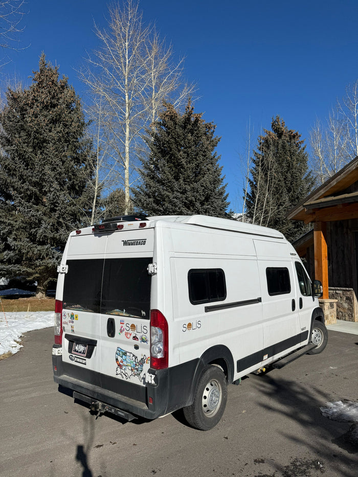 A white Winnebago Solis camper van, popular for rv rental, parked on an asphalt driveway with snow, surrounded by pine and bare trees.