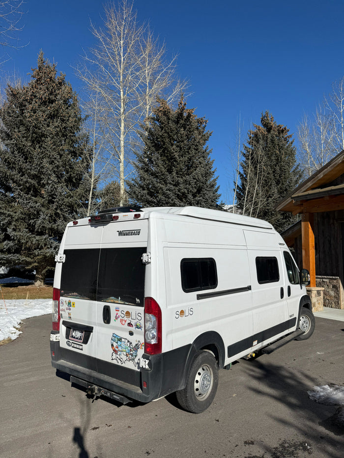 A white Winnebago Solis camper van, popular for rv rental, parked on an asphalt driveway with snow, surrounded by pine and bare trees.