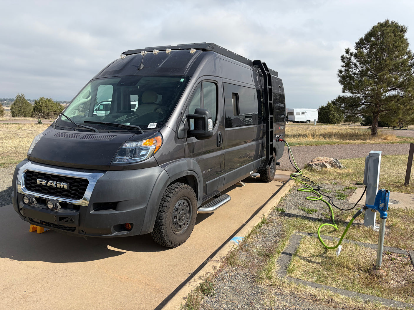 Dark grey RV rental camper van parked at a campsite, hooked up to water and electricity on a cloudy day.