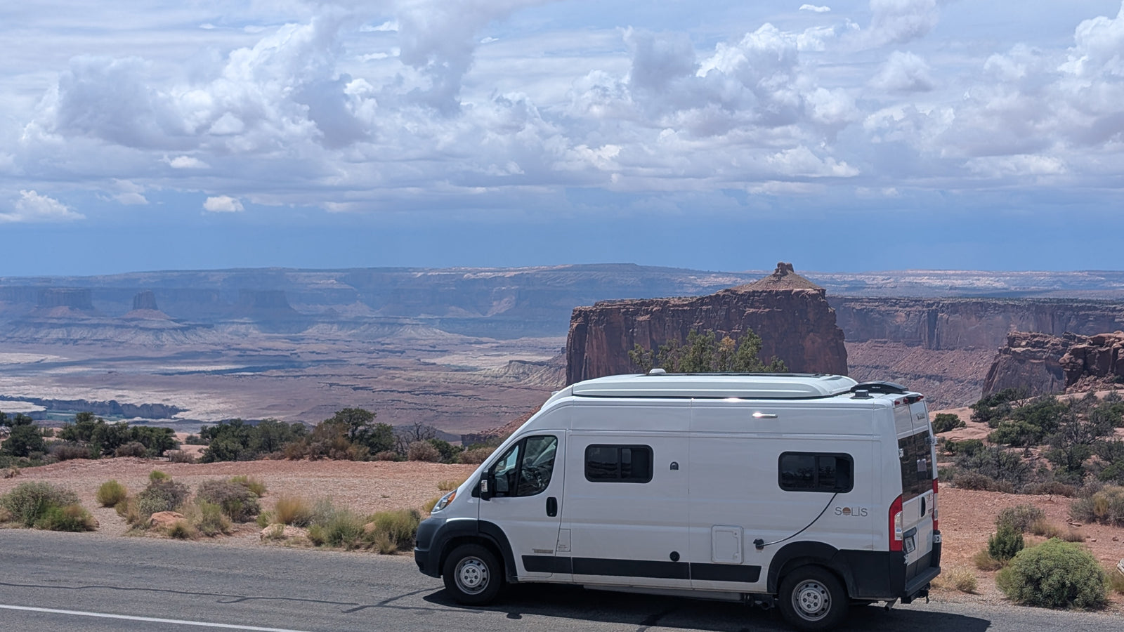 A white rv rental (Winnebago Solis) is parked by a road, offering a panoramic view of layered desert canyons and mesas under a cloudy sky.
