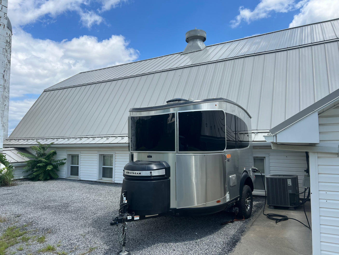 An Airstream RV rental is parked on a gravel driveway next to a barn with a metal roof under a blue sky.