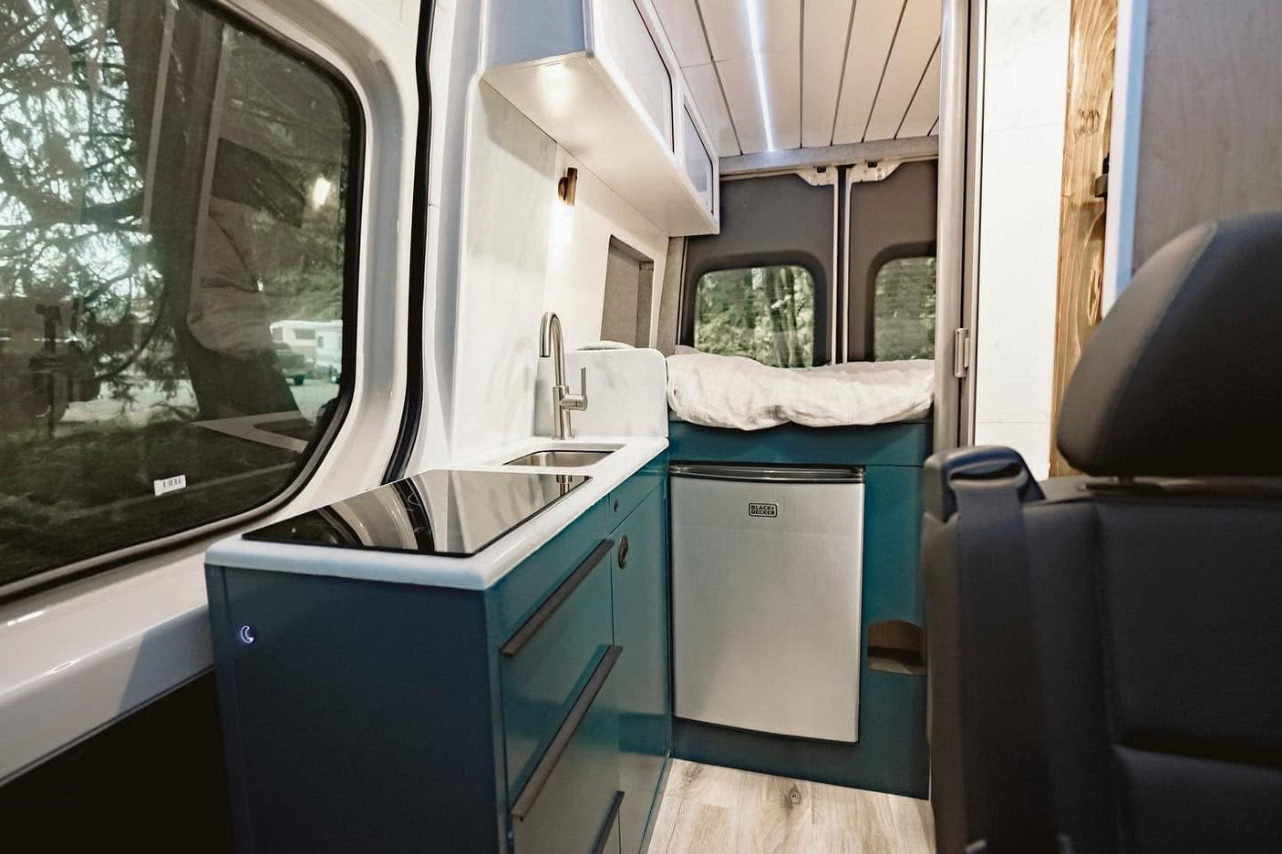 Inside a modern rv rental: compact camper van kitchen with sink, induction cooktop, fridge, and cozy bed in the rear.