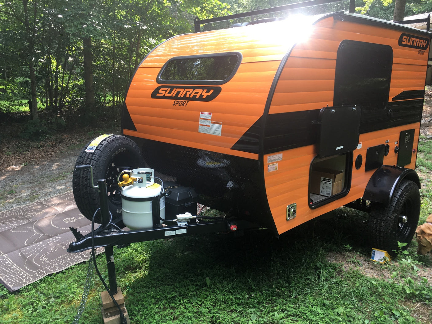 Vibrant orange Sunray Sport teardrop RV rental, with a spare tire & propane tank, parked in a sunny wooded area.