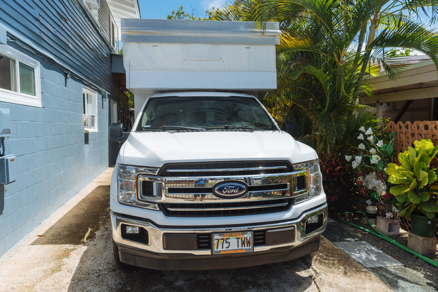 White Ford truck with a camper shell, likely an RV rental, parked on a paved driveway in Hawaii, between a blue house and lush plants.