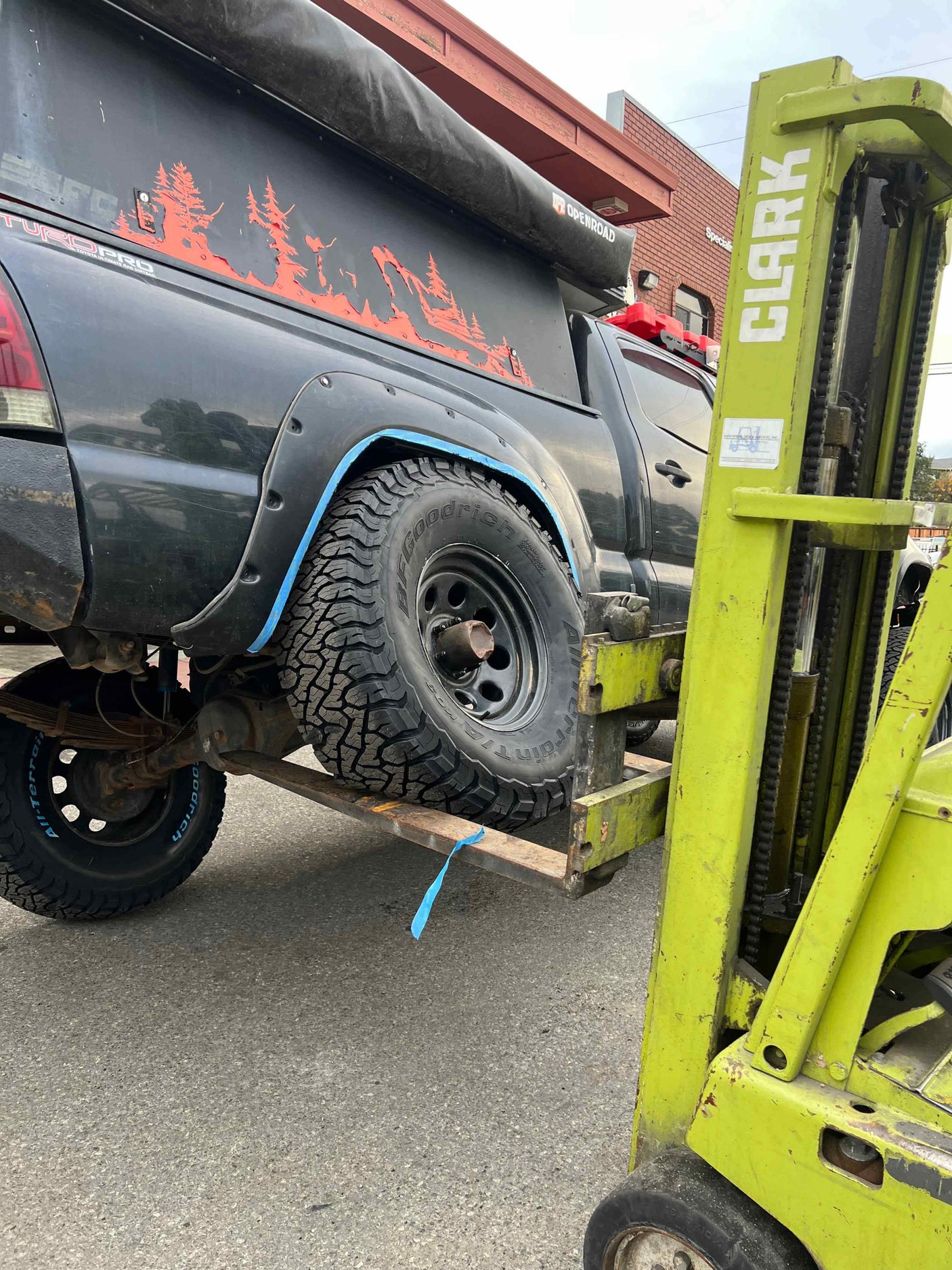 A forklift lifts an off-road truck with a camper, likely for rv rental service or maintenance. Shows BFGoodrich tires.