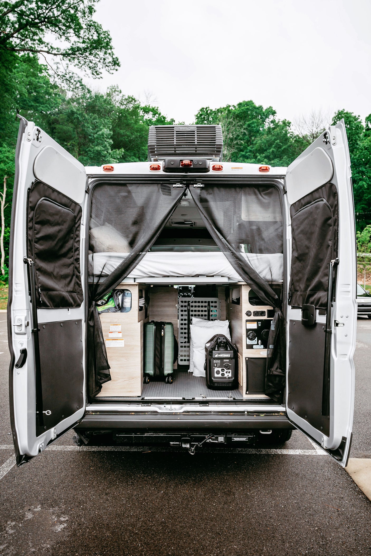 Inside an RV rental camper van: rear doors open, revealing organized storage, a bed, a suitcase, and a Powerhouse generator, ready for adventure.